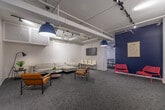 Located at 955 Warwick Court in Burlington, Ontario, this well-appointed single-floor commercial suite offers a thoughtfully designed layout suited for professional or community use. The space is defined by a clean, modern aesthetic, featuring crisp white walls, patterned carpet tiles, and exposed ceiling infrastructure with industrial-style track lighting that adds both function and character.

The main lounge and gathering area is generously proportioned, furnished with a plush sectional sofa, sleek leather accent chairs, and a round coffee table. A bold navy blue accent wall serves as a striking focal point, complemented by framed artwork and vibrant red seating that adds energy to the room. A wall-mounted flat-screen display and additional rows of white chairs suggest the space supports presentations or group meetings.

Adjacent to the lounge is a fully equipped kitchen, finished with floor-to-ceiling white cabinetry, a blue subway tile backsplash, white countertops, large-format tile flooring, and stainless steel appliances including a full-size refrigerator and dishwasher. A glass partition wall elegantly separates the kitchen from surrounding areas while maintaining visual openness.

A secondary, more intimate lounge alcove provides a quieter setting with white textured brick walls and ample seating. Throughout the suite, exposed ductwork and structural elements lend an industrial-modern quality to the overall ambiance.