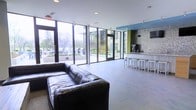 This contemporary communal space is designed with an open-concept layout that maximizes both functionality and visual appeal. Expansive floor-to-ceiling windows line the exterior walls, flooding the interior with abundant natural light and offering clear views of the surrounding outdoor landscaping and neighboring structures. The generous open floor plan creates a welcoming sense of spaciousness throughout.

The lounge area is anchored by a large, plush black leather sectional sofa, providing ample seating for gatherings or relaxed conversation. Smooth, large-format grey tile flooring extends across the entire space, lending a clean and cohesive aesthetic. A standout feature is the built-in bar counter finished in a polished marble-style surface, accompanied by a row of sleek modern bar stools — ideal for casual dining or socializing. Behind the bar, a textured mosaic accent wall adds depth and visual interest, complemented by bold color-blocked architectural elements in green and blue that energize the space.

Recessed ceiling lighting works in tandem with the natural light to ensure the space remains well-lit throughout the day. Access to the outdoor areas is facilitated by multiple glass doors, seamlessly connecting the interior to the exterior environment. The overall ambiance is modern, vibrant, and inviting.
