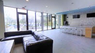 This contemporary communal space is designed with an open-concept layout that maximizes both functionality and visual appeal. Expansive floor-to-ceiling windows line the exterior walls, flooding the interior with abundant natural light and offering clear views of the surrounding outdoor landscaping and neighboring structures. The generous open floor plan creates a welcoming sense of spaciousness throughout.

The lounge area is anchored by a large, plush black leather sectional sofa, providing ample seating for gatherings or relaxed conversation. Smooth, large-format grey tile flooring extends across the entire space, lending a clean and cohesive aesthetic. A standout feature is the built-in bar counter finished in a polished marble-style surface, accompanied by a row of sleek modern bar stools — ideal for casual dining or socializing. Behind the bar, a textured mosaic accent wall adds depth and visual interest, complemented by bold color-blocked architectural elements in green and blue that energize the space.

Recessed ceiling lighting works in tandem with the natural light to ensure the space remains well-lit throughout the day. Access to the outdoor areas is facilitated by multiple glass doors, seamlessly connecting the interior to the exterior environment. The overall ambiance is modern, vibrant, and inviting.
