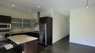 This single-floor space showcases a contemporary open-concept design centered around a well-appointed kitchen. Dark espresso cabinetry provides a striking contrast against the light-toned walls and countertops, creating a bold visual dynamic. The kitchen is equipped with stainless steel appliances, including a full-size refrigerator, and features a gas cooktop complemented by a stainless steel island ventilation hood. Frosted glass upper cabinets on one wall add a modern touch while softening the overall aesthetic. A kitchen island with a light countertop surface offers additional workspace and casual gathering space. Rich, dark hardwood flooring extends throughout, lending warmth and continuity to the open layout. Recessed lighting in the white ceiling provides clean, even illumination across the space. The kitchen flows naturally into an adjacent open area, suggesting a connected living or dining zone. The minimalist design approach, with neutral walls and a carefully curated color palette of dark and light tones, gives the space a refined, modern ambiance suited to contemporary living.