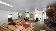 The Club Room is a spacious, single-floor communal space designed to bring residents together in a warm and inviting atmosphere. The open-concept layout seamlessly connects a cozy lounge area, multiple dining spaces, and a kitchenette, making it well-suited for a variety of social gatherings and events.

The lounge area anchors the room with plush brown leather sofas accented by colorful orange and green throw pillows, arranged around a bold, multicolored area rug. A round red leather ottoman serves as a stylish centerpiece, adding a pop of color and functionality to the seating arrangement. A ceiling fan overhead ensures comfortable airflow throughout the space.

Two distinct dining areas are positioned near the windows, each furnished with dark wooden chairs and tables that provide ample seating for groups. Natural light filters in through multiple windows, brightening the interior and creating an open, airy feel. Decorative wall art, including a vibrant tree motif, adds a welcoming touch to the neutral-toned walls.

Toward the back of the room, a kitchenette with white cabinetry and countertops provides convenient access to appliances, making it easy to prepare and serve refreshments. The textured ceiling and carpeted flooring throughout contribute to the room's comfortable and casual ambiance.