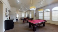 This single-floor amenity facility is thoughtfully designed to offer a variety of recreational and social spaces. The centerpiece is a spacious billiards room featuring two full-size pool tables with rich mahogany finishes and vibrant red felt, illuminated by large yellow drum pendant lights suspended from a coffered ceiling. Arched windows and multiple blinds-covered panes allow natural light to fill the room, while recessed lighting ensures consistent illumination throughout.

Adjacent to the billiards area is a game room equipped with a decorative foosball table and additional seating at a round dining table, offering a more casual setting for group activity. Wall-mounted artwork and panel molding details add a polished aesthetic throughout.

The media lounge features a warm, intimate atmosphere with two-tone walls accented by dark wood trim and crown molding. Multiple wall-mounted flat-screen televisions are paired with comfortable seating, including a cushioned sofa and upholstered armchair, arranged around a round coffee table.

A separate dining nook provides a quiet retreat with a round wood table, white upholstered chairs, and generous window light. Together, these areas create a well-rounded, socially oriented amenity space with consistent, refined finishes.