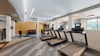 This expansive fitness center is thoughtfully designed to accommodate a wide range of workout styles, organized across several distinct zones that flow seamlessly into one another. The cardio area features a row of modern treadmills positioned along large windows that invite abundant natural light, set atop striped carpet tiles that lend a contemporary feel to the space. Overhead, linear LED lighting runs across smooth ceilings, ensuring bright, even illumination throughout.

The strength training zone is equipped with cable machines, resistance equipment, and a full rack of dumbbells, complemented by wall-mounted mirrors that enhance the sense of openness. A dedicated functional training corridor stands out with its rich warm-toned wood ceiling, turf flooring, and an impressive array of equipment including battle ropes, medicine balls, plyo boxes, a prowler sled, and a large tire — ideal for high-intensity training.

Glass-walled studio spaces provide dedicated areas for cycling and group fitness, maintaining visual connectivity with the rest of the facility while offering acoustic separation. The studios feature wood-plank flooring, ceiling fans, and generous window views of lush greenery outside. Throughout the fitness center, the design balances industrial elements with warm finishes, creating an energizing yet inviting environment for all levels of fitness enthusiasts.