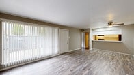 This single-story space is designed with a clean, functional layout that flows logically from room to room. The living area serves as the central hub, featuring wood-look plank flooring in a warm gray tone, neutral taupe-painted walls, and a ceiling fan overhead. A full-width vertical blind treatment frames a large sliding glass door, allowing generous natural light to fill the space. A raised countertop pass-through connects the living area to the kitchen, providing an open sightline between the two spaces.

The galley-style kitchen is efficiently organized with white cabinetry lining both walls, offering ample storage above and below the counters. A stainless steel double-basin sink, electric range, dishwasher, and refrigerator complete the kitchen's functional setup. The wood-look flooring continues throughout, maintaining visual consistency across the unit.

The bedroom areas are comfortably proportioned, each featuring the same neutral color palette, wood-look flooring, and standard closet access. One bedroom includes a window that brings in natural light, while another features two doorways providing access to a closet and a hallway. The bathroom is straightforward in design, equipped with a single-sink vanity, toilet, and a tub-shower combination with chrome fixtures.