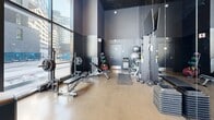 Located at 205 Avenue De l'Équinoxe in Brossard, Québec, this ground-floor amenity space at Lümeo offers a seamlessly integrated lobby and fitness centre with a distinctly contemporary architectural character. The entrance lobby immediately impresses with soaring floor-to-ceiling glazing that floods the interior with natural light and frames views of the surrounding urban landscape. A striking dark slatted ceiling runs throughout, complemented by polished concrete flooring and sleek black structural columns clad in reflective panels that add depth and visual interest to the space. The lounge area is thoughtfully arranged with bold orange upholstered seating and a separate white sofa grouping, divided by a textured three-dimensional feature wall that serves as an elegant focal point. Lush indoor palms and tropical plants add warmth and a sense of vitality to the refined, modern aesthetic. Adjacent to the lobby, the fitness centre is equally impressive, bathed in natural light through expansive glass walls that offer open city views. The gym is well-appointed with a full range of cardio equipment including ellipticals, stationary bikes, and treadmills, alongside a dedicated strength training zone featuring free weights, barbells, benches, medicine balls, and resistance machines. Full-length mirrors and dark painted walls in the strength area give it a focused, professional atmosphere. Together, these spaces deliver an elevated amenity experience.