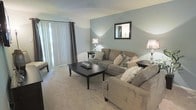 This single-floor space showcases a thoughtfully designed living room that serves as its central feature. The room is finished with soft, neutral-toned carpeting that adds warmth underfoot and complements the cool, muted blue-grey walls, creating a calm and welcoming ambiance throughout.

The seating area is anchored by two generously sized, taupe upholstered sofas arranged around a dark-stained wood coffee table, offering a comfortable and functional layout for relaxation or entertaining. Decorative throw pillows in coordinating tones add visual interest and texture to the seating arrangement.

A large sliding glass door fitted with vertical blinds and patterned curtain panels lines one wall, allowing natural light to filter into the space while providing privacy options. The natural light enhances the room's airy feel.

Additional touches include elegant table lamps with classic bases that provide warm ambient lighting, a framed wall mirror that visually expands the space, and tasteful décor accessories on the coffee table. The clean lines and cohesive color palette give the living area a polished, move-in-ready appearance.