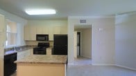 This single-floor residence offers a practical and well-organized layout designed for comfortable everyday living. The kitchen serves as a central hub of the home, equipped with black appliances including a refrigerator, electric range, and built-in microwave. White cabinetry lines the walls, providing ample storage space, while granite-style laminate countertops offer generous prep area. A breakfast bar extends from the counter, creating a natural transition between the kitchen and the adjacent living area, which features neutral carpeting that adds warmth underfoot. The open sightlines between these spaces enhance the sense of openness throughout the unit. A hallway visible from the kitchen leads to a dedicated laundry area, where a stacked washer and dryer unit is conveniently housed. The interior finishes are consistent throughout, with light-toned walls, white trim, and a neutral color palette that lends a clean, cohesive aesthetic. Overhead lighting in the kitchen ensures a bright workspace, while HVAC vents are strategically placed for efficient climate control. The overall layout is thoughtfully designed to maximize functionality while maintaining an inviting and comfortable atmosphere.