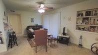 This single-story space centers around a comfortable living room that offers a relaxed, functional layout. The room features wall-to-wall carpeting in a neutral tone, contributing to a warm and quiet atmosphere. A ceiling fan with an integrated light fixture is positioned centrally overhead, providing both air circulation and ambient lighting.

The living area is furnished with a dark leather sofa accented by a decorative pillow, a patterned upholstered armchair, and a small round side table. A traditional wooden media console holds a flat-screen television, while a decorative area rug anchors the seating arrangement. Wall décor includes a motivational sign and a small framed piece, adding personality to the neutral-toned walls.

A large vertical blind window treatment occupies a prominent portion of one wall, suggesting access to natural light and potentially a door or sizeable window beyond. The overall ambiance is casual and lived-in, with a straightforward arrangement that maximizes usable floor space. The single-floor layout offers convenient, step-free accessibility throughout the home.