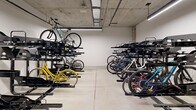 Located at 10401 Motor City Dr in Bethesda, Maryland, this dedicated bicycle storage room offers a well-organized, single-level space designed to efficiently accommodate a large number of bikes. The room features multiple rows of black double-tiered vertical bike racks, maximizing storage capacity by allowing bicycles to be stored both at ground level and elevated on upper tiers. The flooring consists of a smooth, polished concrete surface, while the exposed concrete ceiling houses visible utility piping and bright linear LED lighting that evenly illuminates the entire space. The walls are finished in clean white paint, contributing to a bright and functional environment. A standard access door is visible at one end, providing straightforward entry and exit. The open layout between rack rows allows for comfortable maneuvering when retrieving or storing bicycles. The overall design is utilitarian and purposeful, prioritizing storage efficiency and ease of access. This space serves as a practical amenity for cyclists, offering a secure and organized indoor environment to store bikes safely.