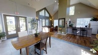 This single-floor space makes a strong architectural impression with its dramatic vaulted ceiling that peaks at a sharp gable, creating a sense of grandeur and openness throughout the interior. The open-concept layout connects a dining area, a casual lounge seating zone, and a standing-height reception or bar counter, allowing for flexible use of the space.

The dining area features a substantial wooden table with matching chairs, positioned atop light-colored tile flooring. This transitions seamlessly into warm hardwood flooring that extends through the lounge and reception areas. The walls are painted in a rich olive-green tone, adding depth and warmth to the interior, while decorative wall accents and sconce lighting contribute to the inviting ambiance.

Natural light is a defining feature of this space. Large windows fitted with blinds line multiple walls, including a striking floor-to-ceiling gabled window arrangement that draws the eye upward and floods the interior with daylight. Additional windows along the side walls and near the entrance further enhance the brightness and provide views of the surrounding greenery.

The lounge area includes comfortable seating and potted plants, adding a welcoming, lived-in feel. A large mirror near the reception area and a tall bar-style counter with stools complete the layout, making this space well-suited for community gatherings, meetings, or hospitality use.