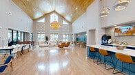 Located at 4000 Galantis Dr in Morehead City, NC, this single-floor community clubhouse offers an expansive, thoughtfully designed gathering space for residents. The centerpiece is a dramatic great room with soaring vaulted ceilings clad in warm wood paneling, oversized cross-pane windows that flood the space with natural light, and stylish woven pendant chandeliers that add a coastal charm. Light hardwood flooring flows throughout, unifying the open-concept layout.

The main lounge area features ample seating with a mix of plush sofas and accent chairs arranged around a central rug, while a teal-accented bar and kitchen island — topped with white countertops and served by sculptural metallic pendant lights — provides a casual café-style hub complete with a coffee station and wall-mounted screens. A teal banquette nook with woven rattan seating adds an intimate dining corner nearby.

A dedicated game and activity zone includes a foosball table and communal dining table, positioned adjacent to a glass-walled fitness center fully equipped with cardio machines and strength training equipment. A secondary lounge area offers built-in wood shelving and large windows overlooking the pool courtyard. The covered outdoor porch, finished in bold blue board-and-batten siding, provides additional seating with views of the surrounding community.