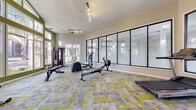 Located at 6147 Winged Elm Ct in Charlotte, NC, this fitness center is a thoughtfully designed, single-floor facility built to support a comprehensive workout experience. The space features soaring vaulted ceilings accentuated by bold green accent walls, which lend an energizing atmosphere throughout. Large windows on multiple walls flood the interior with natural light and offer pleasant views of the surrounding outdoor greenery, creating an inviting and motivating environment.

The flooring consists of vibrant geometric-patterned carpet tiles in shades of gray, white, and yellow-green, adding a modern, dynamic aesthetic to the space. Equipment is well-distributed and includes rowing machines, elliptical trainers, Life Fitness treadmills, cable and multi-use weight machines, adjustable dumbbells with a dedicated rack, and multiple weight benches. A large floor-to-ceiling glass partition wall separates zones within the facility, maximizing openness while defining workout areas.

Wall-mounted flat-screen televisions provide entertainment during workouts, and motivational wall decals, including a bold "NO EXCUSES" statement, reinforce an active mindset. Track lighting runs along the ceiling to ensure even illumination throughout. Frosted glass interior doors connect to additional areas of the facility. Workout reference charts are displayed on the walls for guidance. Overall, the space combines functional design with an uplifting ambiance ideal for fitness-focused use.