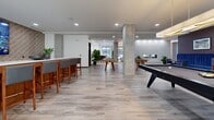 Located at 2358 Bransford Ave in Nashville, TN, this contemporary amenity lounge is a generously proportioned, single-floor social space designed for recreation and community gathering. The open-plan layout is anchored by light gray wood-look flooring that flows seamlessly throughout, complemented by recessed lighting and a clean, modern aesthetic.

The game area serves as the focal point of the space, featuring a sleek black pool table beneath a modern linear pendant light and a foosball table positioned nearby. Floor-to-ceiling glass doors and windows flood the space with natural light while offering views of the surrounding Nashville skyline.

A bar counter with upholstered high-top seating provides a casual gathering spot, while a separate lounge-dining area is defined by a richly textured, deep burgundy accent wall adorned with framed sports photography. Tufted navy blue banquette seating lines the wall, paired with warm wood high-top tables and chairs, creating an inviting, restaurant-quality dining nook. A patterned area rug with golden accents delineates this zone from the wider open floor.

Additional touches include a built-in fireplace feature, lush potted greenery, and a large-screen display mounted to an exposed brick-style wall. Concrete structural columns add an industrial contrast to the otherwise polished interior, giving the space a refined yet approachable character well-suited for social events and everyday relaxation.