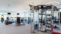 Located at 6650 Liberty Grand Blvd in Powell, Ohio, this modern fitness center offers a spacious, single-floor layout designed to accommodate a comprehensive range of workout activities. The open floor plan spans a generously sized area, providing ample room for members to move freely between equipment zones without feeling crowded.

The space is flooded with natural light through large, floor-to-ceiling and clerestory windows that line the perimeter walls, offering pleasant outdoor views of the surrounding landscape. Striking circular ring pendant lights are suspended from the high, white ceilings throughout, creating a clean and contemporary ambiance.

The facility is organized into distinct functional zones. A dedicated cardio area features rows of treadmills, ellipticals, stair climbers, and stationary bikes — primarily Life Fitness branded — positioned facing the expansive windows. A strength training zone houses a wide array of selectorized machines and cable systems, while a free weights area includes a full dumbbell rack with floor-to-ceiling mirrors. A functional training zone features a multi-station rig with TRX suspension trainers, battle ropes, and a heavy bag.

Accent walls of natural wood and decorative stone tile provide warmth and texture, balancing the otherwise crisp white walls and light wood-tone flooring. Wall-mounted televisions are positioned throughout the space for entertainment during workouts.