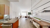 This single-floor residence is defined by its open-concept design and striking architectural proportions, with notably high ceilings that amplify the sense of space across the main living areas. Light wood-look flooring runs continuously throughout, unifying the layout and contributing to a warm, cohesive aesthetic.

The kitchen is well-appointed with rich warm-toned wood cabinetry, stainless steel appliances including a professional-style range and French door refrigerator, a stainless steel ventilation hood, subway tile backsplash, and a grey composite countertop that extends to a peninsula with a double sink—offering both functionality and a natural transition into the dining area.

The dining space is anchored by a solid wood table beneath globe pendant lighting, with a large-scale abstract artwork adorning the adjacent wall. The living room benefits from tall ceilings and sheer floor-to-ceiling drapery that diffuses abundant natural light, creating a serene, airy atmosphere.

The bedroom features dramatic ceiling height, wood flooring, and direct access to a bathroom with warm wood cabinetry, a wide vanity mirror, and a glass-enclosed shower. The overall ambiance throughout is calm, modern, and refined.