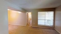 This single-floor space presents a straightforward and functional layout. The main living area is finished with wall-to-wall carpeting in a warm beige tone, providing a comfortable underfoot feel throughout. The walls are painted in a neutral taupe shade, creating a calm and understated atmosphere that offers a versatile backdrop for furnishing and décor.

The textured popcorn ceiling adds a classic, traditional character to the space. A large window fitted with horizontal blinds allows natural light to filter into the room, brightening the interior while offering privacy control. The window overlooks an exterior area, contributing a sense of openness to the otherwise enclosed space.

An open doorway leads from the main living area into an adjoining hallway, which appears to connect to additional rooms deeper within the unit. The hallway is illuminated by interior lighting, suggesting further living spaces beyond. White door trim and baseboards provide clean contrast against the neutral wall color, lending a tidy, well-defined structure to the overall layout. The space is practical and move-in ready, suited for a range of configurations.