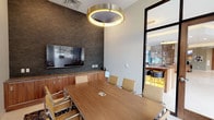This professionally designed conference room offers a polished and functional environment ideal for meetings and presentations. The centerpiece of the space is a substantial wood-grain conference table surrounded by sleek, tan leather chairs with chrome frames, providing comfortable seating for multiple attendees. A large wall-mounted flat-screen display is set against a dramatic textured dark stone accent wall, flanked by warm walnut built-in cabinetry beneath — a combination that delivers both practicality and visual impact. Overhead, a distinctive circular gold pendant light fixture serves as an architectural focal point, casting a warm ambient glow throughout the room. The light-toned tile flooring is complemented by a patterned area rug beneath the table, adding texture and dimension to the space. Floor-to-ceiling black-framed glass walls and a wood-framed glass door open the room to an adjacent lounge area and flood the space with natural light. These expansive windows also frame views of an outdoor courtyard with seating, landscaping, and a surrounding multi-story building. The broader facility visible through the glass entry features a well-appointed lounge with marble accents, decorative partitions, and soft seating, reflecting a high-end, modern commercial interior design throughout.