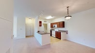 This single-floor residence is designed with a practical, open layout that connects the kitchen and living areas seamlessly. The kitchen is well-appointed with rich, dark wood cabinetry, granite-look countertops, white appliances including a range, microwave, and refrigerator, and tile flooring. A peninsula with a built-in sink and dark countertop extends into the living space, providing additional prep space and a natural division between the kitchen and the carpeted living area. A decorative pendant light hangs above the peninsula, complementing the track lighting overhead.

The living area is generously proportioned, with soft carpet, neutral white walls, and a set of multi-pane windows that allow natural light to fill the room. Built-in shelving is recessed into one wall, offering functional display or storage space. An entry corridor leads from the front door into the main living space, providing a clear transition zone.

The bedroom features plush carpeting, a ceiling fan with light fixture, and a wide window framing an outdoor view, creating a bright and comfortable retreat. A closet with bifold doors provides ample storage. The full bathroom includes a dark wood vanity with a granite-look countertop, a large mirror, tile flooring, and a tub-shower combination. A dedicated laundry area houses a top-loading washer and dryer unit beneath wire shelving.