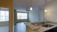 This single-floor space showcases an open-concept layout that seamlessly connects the kitchen and living area. The kitchen features speckled granite countertops with an undermount sink, a peninsula that doubles as a breakfast bar, and a pendant light that adds a warm, focused glow over the workspace. A ceiling-mounted HVAC vent ensures efficient climate control throughout.

The living area extends naturally from the kitchen, finished with durable hardwood-style flooring in a warm, medium-toned hue that runs throughout the space. A ceiling fan with an integrated light fixture serves as a functional centerpiece, promoting air circulation and supplemental lighting. Neutral sage-green walls and white trim create a clean, cohesive aesthetic that complements the natural light streaming through a large picture window. The window frames views of the surrounding neighborhood, enhancing the sense of openness. Subtle architectural details, including raised-panel doors and consistent base molding, contribute to a polished, well-finished interior throughout the space.