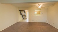 This two-story residence offers a straightforward and functional layout designed for comfortable everyday living. The main level centers around a spacious carpeted living area, providing a warm and inviting atmosphere underfoot. A ceiling fan with an integrated light fixture serves as a practical focal point overhead, supporting air circulation throughout the space. A striking open staircase with metal railings connects the two floors, contributing a sleek, modern element to the interior while maintaining an open, airy feel in the main living space. Adjacent to the living area, a pass-through window opens directly into the kitchen, allowing for easy interaction between the two spaces. The kitchen features wooden cabinetry and standard appliances visible through the pass-through, reflecting a classic and efficient design. The lower level transitions smoothly between carpeted and hard-surface flooring areas, delineating the different functional zones. A floor vent ensures consistent climate control throughout the home. Natural light filters in through a door visible at the base of the staircase, adding brightness to the interior. Overall, the space presents a clean, practical design well-suited for daily residential use across both its levels.