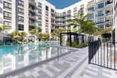 Located at 501 NW Seventh Ave in Fort Lauderdale, FL, this outdoor amenity space showcases a resort-style pool courtyard designed for relaxation and leisure. The centerpiece is a generously sized swimming pool with crystal-clear blue water, framed by sleek tiled pool decking in a sophisticated gray-and-white geometric pattern. Surrounding the pool, abundant tropical landscaping — including tall swaying palms, lush shrubs, and colorful plantings — creates a vibrant, resort-like atmosphere that reflects South Florida's natural character. A series of modern pergola structures with dark metal framing provide shaded seating areas outfitted with wicker lounge furniture, offering an inviting retreat from the sun. Rows of teal and aqua lounge chairs are positioned poolside beneath shade umbrellas for additional outdoor seating. The courtyard is enclosed by a sleek black iron safety fence and is flanked on multiple sides by a multi-story contemporary building with white facades, large windows, and private balconies that overlook the pool below. Glass-paneled entryways connect the outdoor space to the building's interior. The overall ambiance is modern, tropical, and refined — a well-appointed outdoor escape in the heart of Fort Lauderdale.