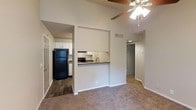 This single-story residence showcases a thoughtfully designed open layout that connects the living area to the kitchen through a convenient pass-through counter. The living space features vaulted ceilings that lend a sense of openness and volume, complemented by a ceiling fan with integrated lighting that provides both comfort and illumination. The flooring transitions between plush carpet in the living area and durable hardwood-style flooring in the kitchen, clearly delineating the two functional spaces.

The kitchen is equipped with white cabinetry providing ample storage, a black refrigerator, a black range, and a single-basin sink positioned at the pass-through counter. The pass-through design enhances connectivity between the kitchen and living area, making the layout feel cohesive and functional. White cabinetry lines the upper and lower walls, maintaining a clean, consistent aesthetic throughout the kitchen.

A hallway extends from the main living area, leading to additional rooms within the unit. The neutral color palette of warm beige and white tones throughout the space creates a clean, welcoming ambiance. The overall design balances practicality with an open, airy feel suited to everyday living.