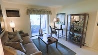 This single-floor residence offers a thoughtfully designed living area that serves as the centerpiece of the space. The living room is bathed in natural light, courtesy of a large sliding glass door dressed with decorative valance curtains, providing direct views of and access to an outdoor patio area with surrounding greenery. The bright, neutral-toned walls and light carpeting enhance the airy feel of the room.

The space is furnished to balance both comfort and style. A plush sofa anchors the seating area, complemented by a dark wood rectangular coffee table set atop a area rug. Along the wall, a sleek console table hosts a classic table lamp and decorative artwork above, adding a refined focal point to the room. A dark wood display cabinet with glass panels stands nearby, offering both storage and an opportunity to showcase decorative items.

A baseboard heating unit runs along the exterior wall beneath the sliding door, a practical feature seamlessly integrated into the layout. Overall, the space conveys a warm, welcoming ambiance with a traditional aesthetic, making it a comfortable and functional living environment.