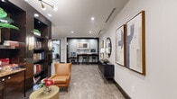 Located at 1900 Yorktown St in Houston's Galleria area, this well-appointed amenity space offers a thoughtfully designed single-floor layout encompassing a living room, dining area, business center, and conference room. The living area features large-format stone-look tile flooring, a wall-mounted flat-screen TV, stylish lounge seating, and a built-in beverage station framed by mosaic tile backsplash — ideal for casual gatherings or relaxed downtime. Adjacent to this is a cozy lounge nook with a sectional sofa, accent chairs in muted tones, colorful throw pillows, and a mirrored wall that enhances the sense of depth and light. The dining area is anchored by a round table with wraparound bench seating beneath a modern sputnik chandelier, creating an inviting setting for communal meals. The business center provides a dedicated workspace with a long wooden desk, multiple computer workstations, and printer access, all framed by dark architectural trim and curated wall art. A private conference room rounds out the space, featuring a large walnut-toned table, ergonomic executive chairs, a tray ceiling with a contemporary globe chandelier, and windows overlooking lush outdoor greenery. Neutral grey walls, recessed lighting, and consistent tile flooring unify the space with a clean, modern aesthetic throughout.