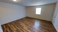This single-floor space presents a straightforward and functional layout with a clean, move-in-ready aesthetic. The interior is defined by rich, dark hardwood-style plank flooring that runs throughout, adding warmth and a sense of continuity to the space. The walls are finished in a soft, neutral tone that pairs well with the flooring and creates a bright, open atmosphere. A window fitted with white blinds allows natural light to filter into the room, enhancing the airy feel of the interior. Dark baseboards provide a subtle contrast along the perimeter, adding a finishing touch to the overall design. The ceiling is smooth and unadorned, keeping the aesthetic simple and versatile. The open, unobstructed floor plan offers flexibility for a variety of furniture arrangements and uses. Overall, the space combines practical design with clean finishes, making it well-suited for a range of occupants and purposes.