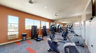 Located at 10435 Lindley Ave in Los Angeles, CA, this fitness center offers a comprehensive and well-appointed exercise facility across a single floor. The main exercise room is generously sized, designed to accommodate a wide variety of workout routines with ample space to move freely between stations. Bold terracotta-orange accent walls energize the space, complemented by a patterned carpet featuring orange highlights that define workout zones throughout the floor. Large windows along the walls invite abundant natural light and outdoor views, creating an open and motivating atmosphere. Ceiling fans ensure consistent air circulation throughout the room. A full lineup of cardio equipment — including treadmills, elliptical machines, and a recumbent bike — lines one side of the room, while the strength training area features cable machines, a multi-rack dumbbell station, a weight bench, and medicine balls. Floor-to-ceiling mirrors on one wall visually expand the space and allow users to monitor their form. A water station and exercise balls round out the functional amenities. The dedicated bathroom features warm tile flooring in a diamond pattern, teal accent walls, wainscoting, accessibility grab bars, and a sleek wall-mounted bench, providing a clean and comfortable space for post-workout use.