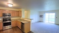 This single-floor space offers a practical and comfortable layout designed for everyday living. The kitchen is a standout feature, outfitted with warm honey-toned wood cabinetry, granite countertops, a stainless steel dishwasher, and a double basin sink. A peninsula counter extends from the kitchen, creating a natural dividing point between the cooking area and the rest of the living space, while also providing additional prep and serving space. The flooring in the kitchen is a rich dark hardwood, adding warmth and contrast to the lighter tones throughout. The adjacent living area transitions to plush carpeting, lending a cozy and inviting feel underfoot. The room is generously proportioned, offering ample space for a variety of furniture arrangements. A window air conditioning unit and a baseboard heating system are both present, ensuring year-round climate comfort. A large window at the far end of the living area allows natural light to fill the space, creating a bright and airy atmosphere. The walls are finished in a soft, neutral tone throughout, providing a clean and versatile backdrop suited to a range of interior styles.