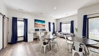 Located at 2745 Campus Pointe Cir in Gainesville, GA, this single-floor amenity space is thoughtfully designed to serve as a multifunctional hub for productivity and community engagement. The interior features crisp white walls, recessed lighting, and wood-look flooring that runs throughout, creating a clean, contemporary aesthetic. Bold navy blue curtains frame multiple large windows fitted with white blinds, allowing ample natural light to fill the room while adding a striking color contrast to the neutral palette. The main area is furnished with multiple round dark-wood tables paired with white metal industrial-style chairs, arranged atop a plush, neutral area rug that defines the central gathering zone. A dedicated computer workstation lines one wall, equipped with multiple monitors and a printer, making it well-suited for academic or professional use. A large whiteboard mounted on the wall provides an additional collaborative tool, ideal for group study or presentations. A cozy lounge area features a light gray sofa accented with decorative pillows, complemented by potted tropical plants that bring a touch of greenery to the space. Vibrant wall art, including a coastal aerial painting and an abstract blue-and-orange canvas, adds personality and visual interest throughout. The overall ambiance is bright, welcoming, and highly functional.