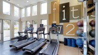 This fitness center is a thoughtfully designed, single-floor space built to accommodate a variety of workout needs. The room immediately draws attention with its soaring ceilings and a generous row of tall windows along one wall, flooding the space with natural light and offering views of the surrounding greenery outside. The bright, open atmosphere creates an energizing environment well-suited for daily exercise routines. A lineup of modern treadmills, each equipped with individual display screens, is positioned to take full advantage of the natural light and outdoor views. On the opposite wall, a bold and vibrant graphic installation featuring large typographic panels in shades of gold, gray, and white adds a dynamic, motivational aesthetic to the space. Exercise mats are neatly stored nearby, indicating provisions for stretching and floor-based workouts. The flooring is durable and fitness-appropriate, complementing the clean, contemporary interior design. Overall, the space balances functionality with a modern visual identity, offering a welcoming and well-lit environment that encourages regular physical activity. The combination of quality equipment, ample space, and thoughtful design details makes this fitness center a standout amenity.