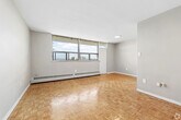 Located at 5-15 Harding Ave in Toronto's Mount Dennis neighbourhood, this 2-bedroom, 1-bathroom unit offers a practical and well-laid-out single-floor design. Classic parquet wood flooring runs throughout the living areas and bedrooms, adding warmth and character to the neutral grey-toned walls. The primary bedroom is a comfortable size, featuring large windows that invite abundant natural light and views of the surrounding greenery, complemented by baseboard heating units along the walls. The second bedroom also features the same parquet flooring and is a well-proportioned space. The kitchen is equipped with ample white cabinetry reaching to the ceiling for generous storage, a marble-patterned countertop, a stainless sink, and white appliances including a stove and refrigerator, all set on light ceramic tile flooring. A sightline from the kitchen opens toward the bright, window-facing living area, enhancing the sense of openness. The bathroom includes a full soaking tub with a shower fixture and cream-tiled surround. Baseboard heating is present throughout the unit, ensuring year-round comfort. Overall, the space presents a clean, functional layout suited to everyday living.