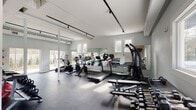The fitness center at 703 Whitney Ave in New Haven, Connecticut is a well-appointed, single-floor exercise facility designed to support a comprehensive workout experience. The space features a clean, modern aesthetic with white walls, white-painted structural columns, and dark rubber flooring that provides both durability and safety underfoot.

The cardio zone is lined with TRUE-brand treadmills, stationary bikes, and cycling equipment, all positioned along a full-length mirrored wall that amplifies the sense of space and allows users to monitor their form. Multiple windows along the walls invite abundant natural light throughout the facility, creating a bright and energizing atmosphere.

The strength and free-weight area is equally well-equipped, featuring a multi-function cable machine, an adjustable workout bench, a tiered dumbbell rack with a full range of weights, and a set of kettlebells and medicine balls neatly organized on dedicated storage racks. A stability ball and medicine ball rack round out the functional training options.

Track lighting mounted along the ceiling provides consistent, focused illumination throughout the space. Glass doors open directly to an outdoor patio area, offering ventilation and a seamless connection to the exterior. The overall layout is open and spacious, allowing for comfortable movement between exercise stations while maintaining a clean, professional environment.