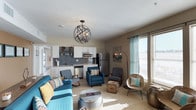 This single-floor residence offers a thoughtfully designed open-concept layout that seamlessly integrates the living and kitchen areas into one cohesive, functional space. The kitchen is outfitted with crisp white cabinetry, a classic subway tile backsplash in a cool gray tone, stainless steel appliances including a refrigerator, dishwasher, and microwave, and ample counter space — making it both practical and visually appealing. A statement orb-style chandelier with exposed candelabra bulbs serves as a striking focal point overhead, adding an industrial-chic touch to the open layout. The living area is generously sized and bathed in natural light streaming through large windows dressed with sheer panel curtains, creating a warm and inviting atmosphere throughout the day. The space is further enhanced by neutral wall tones that complement the varied textures and colors of the furnishings, including teal upholstered seating, woven accent chairs, and eclectic decorative accessories. Soft carpeted flooring adds comfort underfoot, tying the living zone together. The overall ambiance is modern yet relaxed, combining functional design with a curated, welcoming aesthetic well-suited for everyday living.