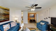 This single-floor residence offers a welcoming and functional open-concept layout that seamlessly connects the main living spaces. The living room is bright and inviting, featuring a ceiling fan with integrated lighting, light-toned flooring, and neutral white walls that provide a clean, airy backdrop. Comfortable seating and tasteful décor create a relaxed atmosphere ideal for everyday living.

A partial dividing wall leads into the dining area, which is distinguished by a warm, deep orange accent wall that adds a bold pop of color and visual interest to the space. A chandelier overhead and a dark wood dining set anchored by a patterned area rug give the dining area a cozy, defined character.

Adjacent to the dining space, the kitchen is visible in the background and features white cabinetry, providing a bright and practical cooking environment. The open sightlines between the living, dining, and kitchen areas contribute to an overall sense of spaciousness and connectivity throughout the home.

Natural light filters through the space, enhancing the warm tones of the décor and the contrast between the neutral and accent-colored walls. The single-floor design ensures easy accessibility and a cohesive flow from one area to the next, making this a practical and comfortable living environment.