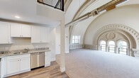 This remarkable two-floor residence occupies a converted space within a historic building, seamlessly blending preserved architectural grandeur with contemporary updates. The main living area is the undeniable centerpiece, featuring dramatically soaring vaulted ceilings adorned with exceptionally detailed ornamental plasterwork. Elaborately carved arches, decorative friezes, and a striking carved medallion surround arched windows that flood the space with natural light, creating an atmosphere of extraordinary character. Exposed ductwork runs along the vault, adding an industrial counterpoint to the classical ornamentation. Soft carpet covers the generous living area floor, complementing the bright, neutral palette throughout.

The kitchen is equipped with white shaker cabinetry, granite countertops, and a full suite of stainless steel appliances, including a side-by-side refrigerator, range, and dishwasher, set atop durable wood-look flooring. A loft-level mezzanine, accessible via an open railing, provides additional functional space above.

The second floor houses additional rooms finished with neutral carpet and clean walls, offering versatile, quieter spaces. The bathroom features a tub-shower combination, white vanity with granite countertop, and wood-look flooring, finished with brushed nickel fixtures throughout.