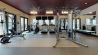 This single-floor amenity center offers a thoughtfully designed collection of spaces catering to both active and professional needs. The fitness facility is the centerpiece, spanning multiple interconnected areas with distinct zones for cardio and strength training. The main gym floor features treadmills, ellipticals, and cable machines beneath a bold deep-red ceiling with large ceiling fans and full-wall mirrors that enhance the sense of space. A dedicated free-weights area houses dumbbell racks and adjustable benches, while a separate functional training room provides hardwood-style flooring and a well-stocked rack of medicine balls, resistance bands, and exercise mats. Large glass doors throughout the fitness area invite natural light and offer views to the exterior. Adjacent to the gym, a stylishly appointed lounge features deep charcoal walls, plush seating, a wall-mounted flat-screen TV, and curated artwork, creating a sophisticated retreat. The conference room accommodates group meetings around a large rectangular table, complemented by wall-mounted display screens and elegant vertical artwork. A business center rounds out the space, offering workstation computers, a printer, granite countertops, and ample desk space beneath a tray ceiling with natural window light.