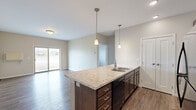 This thoughtfully designed single-floor unit offers 2 bedrooms and 1 bathroom within a comfortable, efficiently laid-out floor plan. The open-concept kitchen and living area forms the heart of the home, anchored by a large center island with granite-style countertops, an integrated sink, and pendant lighting overhead. Dark wood cabinetry, recessed lighting, and black appliances—including a refrigerator, range, and microwave—complete the kitchen. The living room extends naturally from this space, featuring dark hardwood-style flooring and a sliding glass door that fills the room with natural light and provides outdoor views. Both bedrooms are carpeted and finished with neutral tones, offering a calm and comfortable atmosphere; the primary bedroom provides a generous amount of space. The full bathroom includes a tub-shower combo, a vanity with matching granite-style countertops, dark wood cabinetry, and wood-look flooring. A dedicated in-unit laundry room houses a full-size washer and dryer. A closet provides additional storage. Throughout, neutral wall tones, consistent flooring materials, and clean white trim contribute to a cohesive, modern aesthetic.