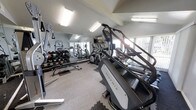 This fitness center offers a thorough and well-organized workout environment housed within a single-floor facility. The space is outfitted with a diverse selection of professional-grade equipment, including Matrix stair climbers, elliptical machines, and treadmills for cardiovascular training. A dedicated free weights area features a full rack of dumbbells alongside a flat bench, catering to strength training needs. The room is illuminated by overhead fluorescent lighting, supplemented by natural light streaming in through large windows fitted with sheer curtains, creating a bright and energizing atmosphere. Full-length mirrors line one wall, visually expanding the space and allowing users to monitor their form during workouts. The flooring combines carpet with interlocking foam mat sections in the free weights area for added comfort and safety. The light gray walls and clean, neutral color palette contribute to a focused and motivating environment. The thoughtful layout ensures ample space between equipment, allowing for comfortable movement throughout the facility. Overall, this fitness center provides a practical, well-lit, and fully equipped setting suitable for a wide range of fitness goals.