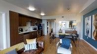 This well-appointed single-floor residence offers a thoughtfully designed open-concept layout that seamlessly connects the kitchen, dining, and living areas. The kitchen is outfitted with rich dark wood cabinetry, stainless steel appliances including a refrigerator, range, and microwave, and a double sink with ample counter space. A second kitchen configuration features sleek gray cabinetry paired with modern appliances, offering a clean, contemporary aesthetic. Warm hardwood flooring runs throughout the space, lending a cohesive and inviting feel. The dining area accommodates a glass-top table with modern chairs, while the living area is furnished with comfortable seating and accented with decorative pillows in yellow and blue tones. Natural light filters in through large windows dressed with sheer white curtains, brightening the living space. A hallway leads to additional rooms, with a stacked washer and dryer unit conveniently tucked behind louvered doors. The overall ambiance is modern and sophisticated, combining functional design with warm, stylish details that create an comfortable and livable environment.