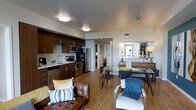 This well-appointed single-floor residence offers a thoughtfully designed open-concept layout that seamlessly connects the kitchen, dining, and living areas. The kitchen is outfitted with rich dark wood cabinetry, stainless steel appliances including a refrigerator, range, and microwave, and a double sink with ample counter space. A second kitchen configuration features sleek gray cabinetry paired with modern appliances, offering a clean, contemporary aesthetic. Warm hardwood flooring runs throughout the space, lending a cohesive and inviting feel. The dining area accommodates a glass-top table with modern chairs, while the living area is furnished with comfortable seating and accented with decorative pillows in yellow and blue tones. Natural light filters in through large windows dressed with sheer white curtains, brightening the living space. A hallway leads to additional rooms, with a stacked washer and dryer unit conveniently tucked behind louvered doors. The overall ambiance is modern and sophisticated, combining functional design with warm, stylish details that create an comfortable and livable environment.