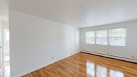 This single-floor unit presents a clean, straightforward layout designed for comfortable everyday living. Polished hardwood flooring flows consistently through the living areas and bedrooms, reflecting natural light and lending warmth to the otherwise neutral, white-painted walls and ceilings. The living room is generously proportioned, offering ample space for furnishings, and benefits from a window that provides views of surrounding greenery and neighboring structures. Baseboard heating units are present throughout, ensuring efficient climate control.

The layout connects seamlessly to a bedroom featuring bifold closet doors that open to a well-sized storage area, along with its own baseboard heating and hardwood floors. A separate room with multiple windows allows for additional natural light, making it well-suited as a second bedroom or flexible living space.

The kitchen is functionally equipped with warm-toned wooden cabinetry, dark countertops, a gas range, over-the-range microwave, and a full-size refrigerator. A window above the sink provides natural light, and durable tile flooring completes the space. The bathroom features a tub-shower combination, a wood-cabinet vanity, light wood-style flooring, and a window for ventilation. A dedicated entryway leads into the unit, providing a clear transition into the main living area.