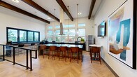Located at Kinect @ Burien in Burien, WA, this single-floor community space combines contemporary design with warm, inviting materials to create a highly functional and visually striking gathering area. Soaring ceilings with prominent dark-stained exposed wood beams establish a bold architectural character, while crisp white walls and abundant natural light maintain a bright, open atmosphere throughout.

The herringbone-patterned hardwood flooring serves as a unifying element across the space, lending a refined yet approachable aesthetic. A fully equipped kitchen anchors one side of the room, featuring dark cabinetry with navy-blue accents, sleek dark marble countertops with dramatic veining, stainless steel appliances, and a wall-mounted range hood. A generously sized island with bar seating accommodates multiple guests, making it ideal for social gatherings and events. Glass pendant lights hang above the island, adding an elegant finishing touch.

Floor-to-ceiling windows line the kitchen wall, flooding the space with natural light and framing panoramic views of the surrounding Burien neighborhood and tree canopy. A wall-mounted television provides additional functionality. The entry area features modern abstract artwork and steel-railed accessible ramp access, while double doors open to an adjacent outdoor terrace, further extending the usable space.