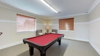 This single-floor space is designed as a recreational and leisure retreat, centered around a well-appointed billiards room. The centerpiece is a full-size pool table with a bold red felt surface and dark wood frame, positioned to allow ample movement on all sides. A cue rack mounted on the wall keeps equipment organized and accessible.

The room's interior design is refined yet comfortable, featuring classic beadboard wainscoting along the lower half of the walls, painted crisp white and topped with a wide chair rail. The upper walls are finished in a warm neutral tone, while elegant crown molding frames the ceiling throughout. Patterned wall-to-wall carpeting in a muted grey adds warmth underfoot.

Natural light filters into the room through multiple windows dressed with warm-toned wood blinds, lending a bright and inviting atmosphere. Overhead, a flush linear light fixture provides consistent illumination across the playing surface.

A dedicated seating nook within the space features two plaid upholstered wingback chairs arranged around a side table with a decorative lamp, offering a relaxed spot for conversation or watching play. A solid wood panel door with classic detailing opens to an adjacent area, further connecting the space. Overall, the room strikes a balance between functional recreation and tasteful, traditional interior design.