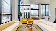 Located at 2171 E 103rd St in Cleveland, Ohio, this single-floor common area space spans a generously sized footprint designed with a contemporary, open aesthetic. The centerpiece is a dramatic lobby-style living room characterized by soaring double-height ceilings and expansive floor-to-ceiling windows that flood the interior with natural light and provide sweeping outdoor views of the surrounding neighborhood. Large-format light tile flooring runs consistently throughout, reinforcing the clean, modern design language.

The living area is furnished with cream sofas, bold mustard-yellow accent chairs, and layered area rugs, creating an inviting and sophisticated atmosphere. Tall white cylindrical columns punctuate the space, complementing the bright, neutral color palette. An adjacent kitchenette features warm natural wood cabinetry, white subway tile backsplash, stainless steel appliances including a full-size refrigerator, and a white central island — offering a fully functional and polished amenity space.

Three hallways connect the common areas, with sage green accent walls adding warmth and color continuity. One hallway leads past glass-partitioned office or conference rooms and a mailroom area equipped with individual mailboxes and elevator access. The overall ambiance is professional yet welcoming, reflecting a well-appointed, modern residential or mixed-use building common area.