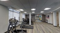This fitness center offers a well-organized and functional workout environment designed to accommodate a range of exercise needs. The space is outfitted with a row of treadmills aligned along one side, providing ample cardio equipment for users. Recessed panel lighting in the drop ceiling ensures bright, even illumination throughout the room, creating a clean and energizing atmosphere.

The flooring features multi-toned, striped luxury vinyl planks that add a contemporary aesthetic while remaining durable and easy to maintain. A central structural column divides portions of the space without significantly disrupting the open layout. Neutral gray walls and white trim keep the environment feeling fresh and professional.

Toward the back of the space, a dedicated stretching and functional fitness area is visible, equipped with colorful exercise balls in various sizes, suggesting options for balance and core training. A framed piece of wall art adds a subtle decorative touch to that area. Natural light filters in through roller-shade windows along one wall, complementing the artificial lighting. Overall, the space is practical, modern, and well-maintained.