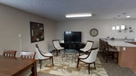 This single-floor space is designed as a comfortable lounge or common area, thoughtfully arranged to encourage relaxation and conversation. The main seating area is anchored by a round glass-top coffee table surrounded by four dark wood-framed chairs upholstered in white fabric, creating an inviting gathering spot. A flat-screen television mounted on a low black media stand serves as a focal point against the neutral-toned walls, making the space well-suited for entertainment. A decorative analog wall clock and a framed geometric mirror add visual interest to the walls, contributing to a warm and homey atmosphere. The flooring features a large area rug with an earthy floral pattern in tones of brown, green, and cream, grounding the seating arrangement and adding texture underfoot. Overhead, a flush-mounted fluorescent light fixture provides practical illumination beneath a textured popcorn ceiling. To the right side of the space, a partial view of bar-height seating and a countertop suggests an adjacent dining or kitchenette area. The overall ambiance is casual yet well-appointed, offering a functional and welcoming environment.