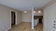 This single-floor space showcases a thoughtfully designed open-concept layout that seamlessly connects the living area and kitchen. Light hardwood flooring runs throughout, lending warmth and continuity to the interior. Neutral beige walls provide a clean, versatile backdrop that complements the overall aesthetic.

The kitchen is a standout feature, equipped with crisp white cabinetry, a stone tile backsplash in earthy tones, and dark countertops that offer a striking contrast. A curved peninsula with integrated open shelving adds both functionality and visual interest, serving as a natural divider between the kitchen and the main living space. A stainless steel dishwasher completes the kitchen's modern appointments.

A structural column near the kitchen peninsula adds an architectural element to the open floor plan. Recessed lighting installed throughout the ceiling ensures even, ambient illumination, while a central pendant light fixture adds a decorative touch to the living area.

Doorways lead to additional rooms, hinting at a thoughtful and well-organized floor plan. Clean white baseboards and trim provide sharp definition throughout the space. The overall ambiance is bright, modern, and move-in ready, making it an inviting and practical environment.
